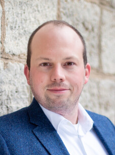 Sebastia Klembt mit Anzug, im Freien vor einer hellen Steinmauer.